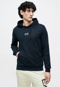 Felpa con cappuccio blu navy con tasca frontale, cappuccio con coulisse e logo EA7 bianco sul petto. Il tessuto sembra liscio ed elastico.