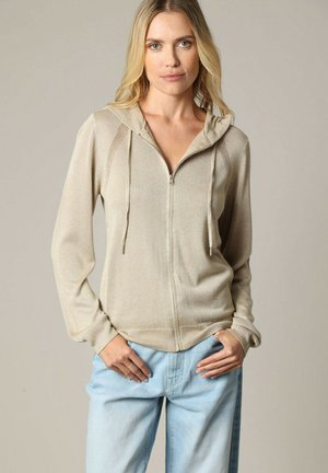 Haut à capuche zippé beige en tissu léger et texturé, avec une capuche à cordon de serrage et des poignets côtelés, associé à un jean en denim bleu clair.