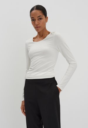 Frau mit dunklem Haar, die ein weißes langärmeliges Oberteil und eine schwarze Hose trägt, mit einer Hand in der Tasche vor einem schlichten hellen Hintergrund steht.