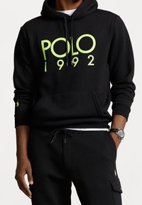 Sudadera con capucha negra de mezcla de algodón, con un gran logo "POLO 1992" en verde lima. Incluye un bolsillo canguro y cordones. Capucha adjunta.