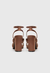 Sandales à talons hauts en satin marron avec un talon épais, une bride de cheville et un détail de nœud décoratif à l'avant. Texture lisse et bords arrondis.
