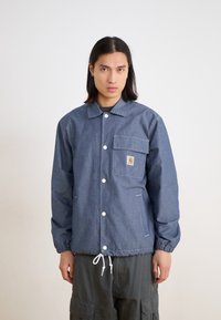 Denim jacket with a button front, chest pocket, and drawstring hem. Features white stitching and a logo patch on the left chest.