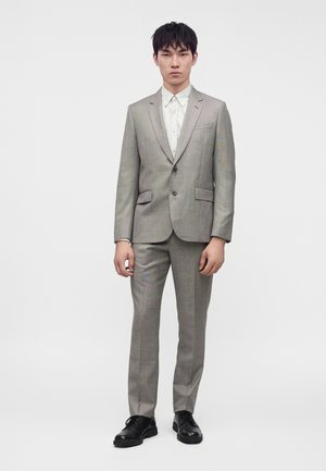 Jeune homme portant un costume à carreaux gris clair, une chemise blanche et des chaussures noires, debout devant un fond blanc uni.