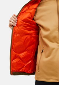 Hellbraune Zip-Jacke mit einer orangen isolierenden Fütterung. Sie hat eine glatte Außenseite und ein strukturiertes Innenmaterial sowie eine Seitentasche.