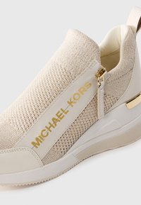 Baskets slip-on beiges avec texture tricotée, lettrage doré "MICHAEL KORS", fermeture éclair sur le côté et semelle en caoutchouc blanc avec une touche dorée.