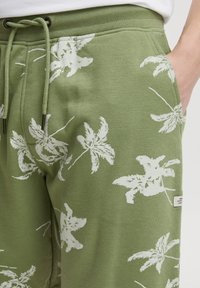 Green cotton joggers with white leafy palm print, drawstring waistband, and side pockets. Tag visible near pocket. Soft texture.