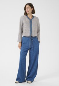 Woman looking right wearing a textured beige and blue jacket with embroidered trim, wide-leg blue jeans, and white shoes on a white background.