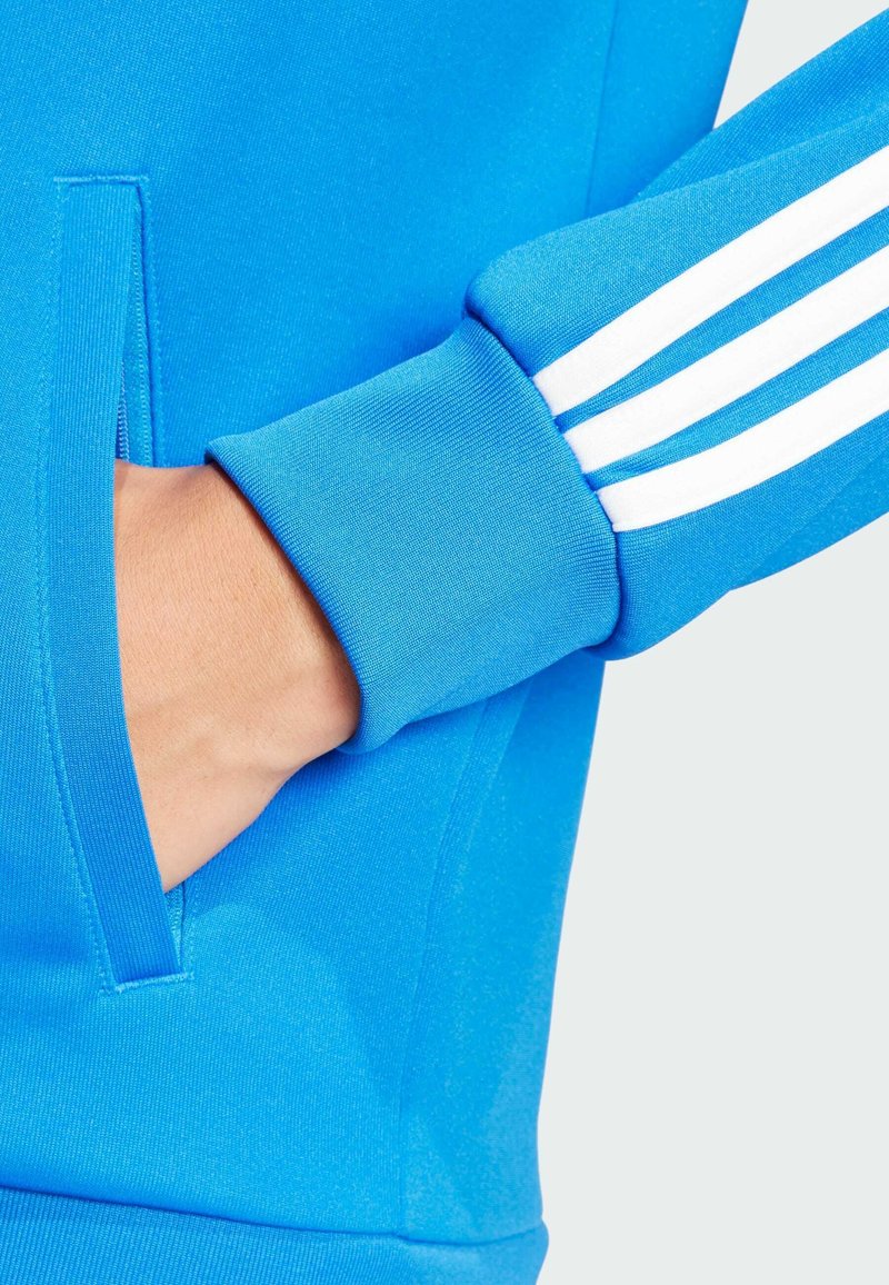 Niebieski sportowy sweatshirt z kieszenią na zamek i trzema białymi paskami na rękawie. Miękka, teksturowana tkanina o dopasowanym kroju.