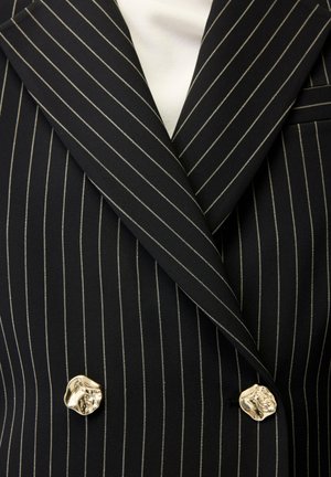 Schwarzer doppelreihiger Blazer mit Nadelstreifen, breiten Revers und unregelmäßigen goldenen Knöpfen über einem weißen Top.