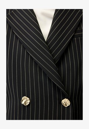 Zwarte dubbelgeknoopte blazer met krijtstreep, brede revers en onregelmatige gouden knopen, gedragen over een wit shirt.