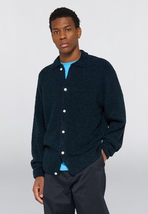 Man draagt een donkerblauwe, knoopjes sluitende, pluizige cardigan over een felblauw overhemd met een donkere broek, staand met één hand in de zak tegen een effen achtergrond.