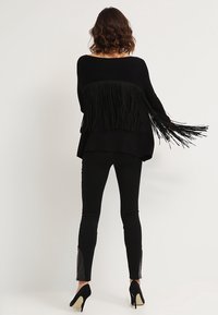 Kvinna sedd bakifrån i en svart fransad tröja, svarta slim fit-byxor med läderlappar och svarta högklackade skor mot en enfärgad bakgrund.