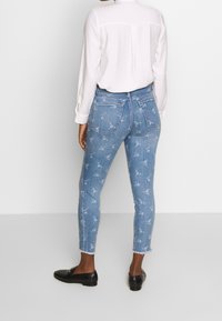Jeans skinny de color azul claro con estampados florales, dobladillo deshilachado y diseño de cinco bolsillos, combinados con una camisa blanca de mangas largas y mocasines negros.