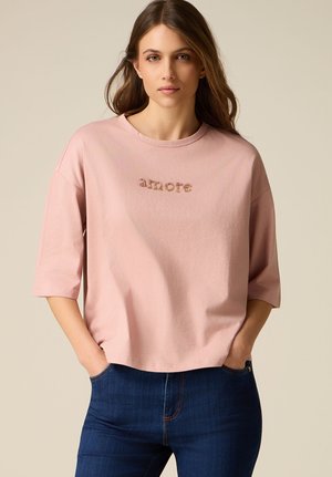 Mujer joven con cabello largo y castaño, viste una camisa rosa suelta con el texto "amore" y jeans azul oscuro, manos en los bolsillos, fondo neutro.