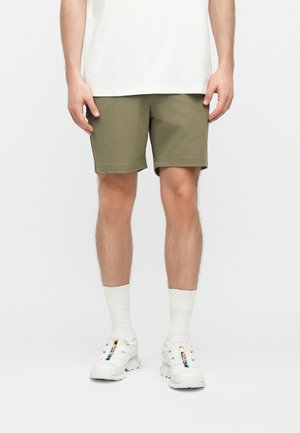 Mees, kellel on seljas oliivirohelised lühikesed püksid, valged keskkõrged sokid, valged tossud musta ja sinise detalidega, seisab tavalise heleda tausta ees.