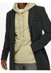 Lange, donkergrijze wollen jas over een lichtbeige hoodie met trekkoorden. De jas heeft een gelieerde reverskraag en een enkele borstzak.