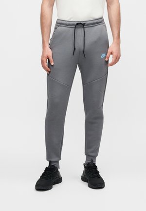 Mężczyzna w szarych spodniach dresowych Nike z niebieskim logo, czarnym sznurkiem i czarnych sneakersach, stojący na tle jednolitego jasnego tła.