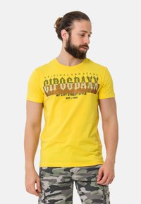 Geel katoenen t-shirt met een ronde hals en een grafische print met tekst en patronen. Gepaard met grijze camouflage shorts.