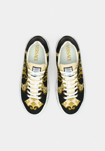 Juodos ir auksinės spalvos sportbačiai su baltais batraiščiais, storu baltu guminiu padu ir ant liežuvėlio esančiu „Versace“ ženklu.