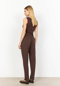 Bruine tanktop en bijpassende joggingsbroek van zachte stof. De broek heeft een elastische tailleband en zijzakken. Platte schoenen met studs.
