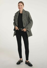 Kobieta stoi uśmiechając się, ubrana w szary płaszcz w jodełkę, czarną bluzkę, czarne spodnie, czarny pasek oraz czarne mokasyny z złotym łańcuszkiem.