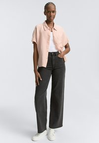 Camicia rosa chiaro a maniche corte con bottoni indossata sopra una maglietta bianca, abbinata a jeans neri a vita alta con gamba larga e sneakers bianche.