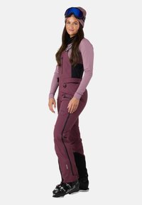 Skioutfit i dyb burgunder med sorte accenter, har et tætsiddende design, justerbare stropper, flere lynlås-lommer og er parret med sorte skistøvler.
