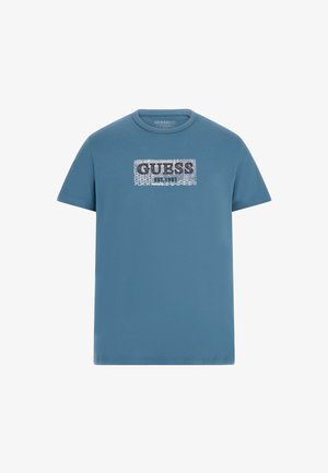 T-shirt in cotone blu con una grafica rettangolare che presenta la parola "GUESS" in lettere grasse argentate, e "EST. 1981" sotto in testo più piccolo.