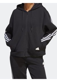 Svart zip-up hoodie tillverkad av mjukt tyg, med vita tre-stripes detaljer på ärmarna och en präglad logotyp på nederkanten. Framfickor ingår.