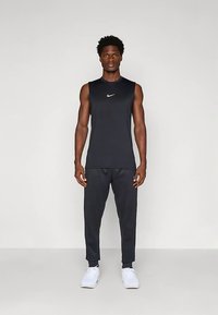 Top sportivo nero senza maniche con logo bianco, abbinato a pantaloni jogger neri coordinati. Modello che indossa scarpe da ginnastica bianche. Design semplice e aderente.