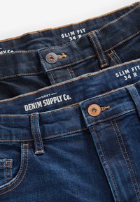 Tummansiniset slim fit -farkut. Varustettu kontrastivärisellä oranssilla ompelulla, metallinappikiinnityksellä ja "Denim Supply Co." -merkinnällä.