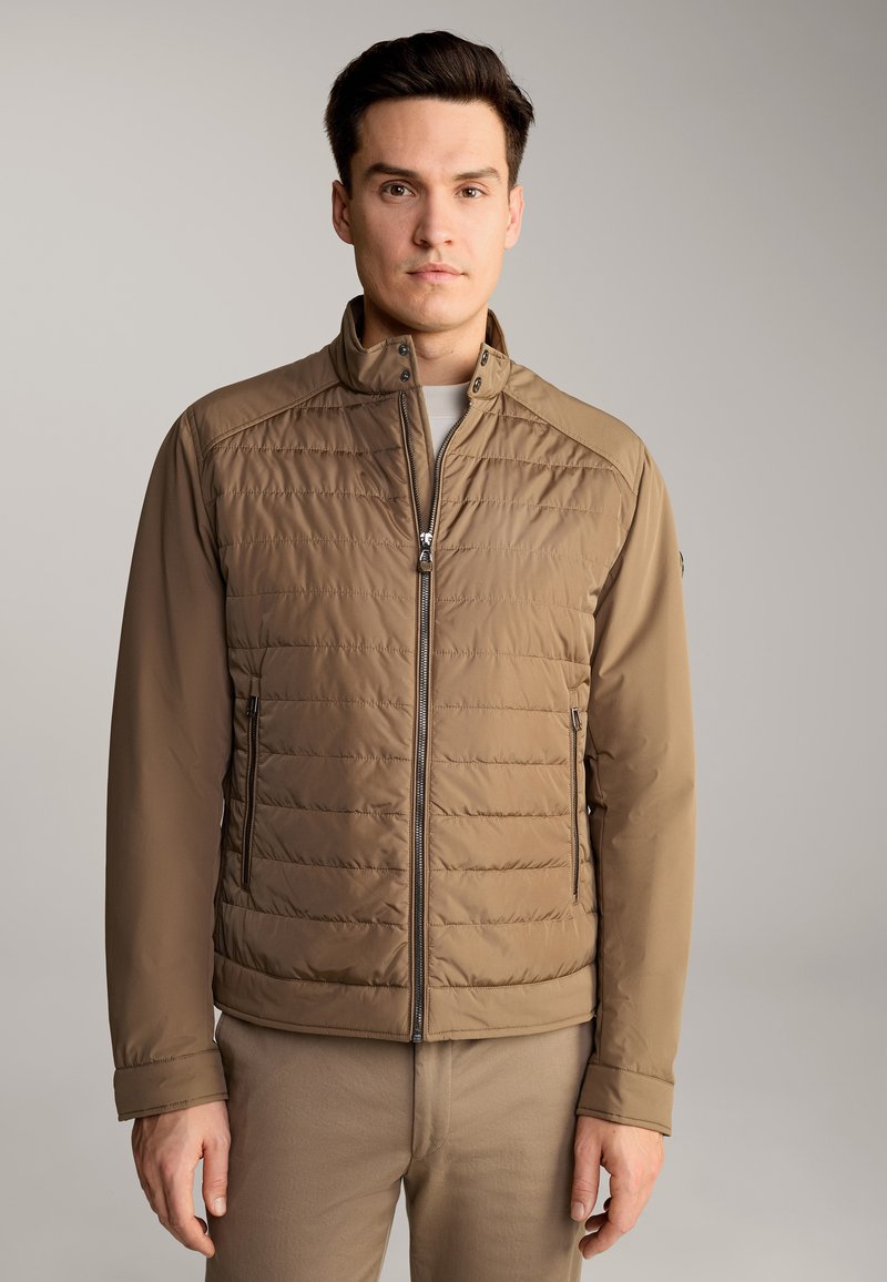 Giacca trapuntata marrone chiaro con colletto alto, chiusura con zip e due tasche laterali. Tessuto morbido con un design leggermente sagomato.