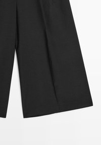 Pantalon noir à jambes larges avec une texture de tissu lisse, présenté de la moitié de la cuisse à l’ourlet sur fond blanc.