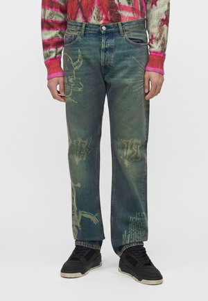 Blaue Herrenjeans mit verblassten Schädel- und Textgrafiken, getragen mit schwarzen Sneakers und einem bunten roten und rosa Pullover.