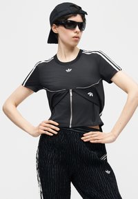 Γυναίκα με μαύρη φόρμα Adidas με λευκές ρίγες, μαύρο καπέλο φοράει ανάποδα, μαύρα γυαλιά ηλίου, με τα χέρια στη μέση μπροστά σε απλό φόντο.