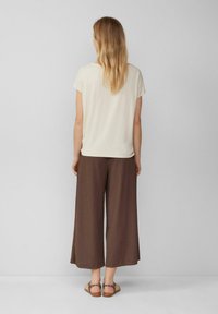 Vrouw met lang blond haar staat met haar gezicht naar een muur, gekleed in een crème top met korte mouwen, bruine wijde broek en sandalen met panterprint.