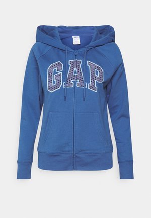 Sweat à capuche zippé bleu en tissu doux, avec un grand logo "GAP" à motif pointillé, des cordons de serrage ajustables et des poches avant.