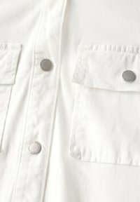 Camicia bianca in cotone con due tasche sul petto dotate di bottoni a pressione in metallo e un tessuto liscio e con texture.