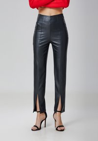 Pantalons en cuir synthétique noir à taille haute, avec des coutures frontales et des fentes jusqu'à la cuisse. Associés à des sandales à talons noirs à lanières.