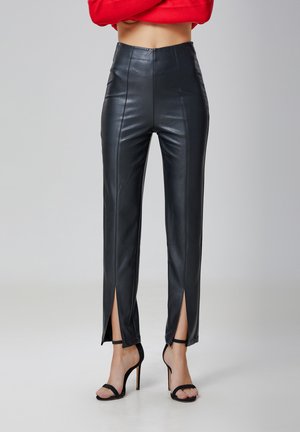 Pantalons en cuir synthétique noir à taille haute, avec des coutures frontales et des fentes jusqu'à la cuisse. Associés à des sandales à talons noirs à lanières.
