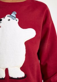 Rotes Sweatshirt mit einem plüschigen weißen Eisbären-Design, verziert mit einer blauen Strickmütze und schwarzer Stickerei, strukturiertem Stoff.