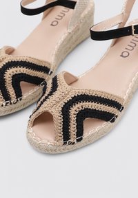 Sandalias de cuña con un diseño trenzado en beige y negro, punta abierta y suela de cuerda de yute con tiras de acento negras.