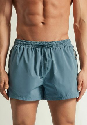 Mandlig overkrop iført blå bade-shorts med elastisk talje, foran snøre og side nøgleclips, stående med afslappede arme.