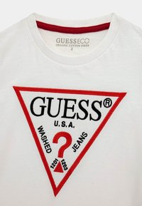 Biała koszulka wykonana z organicznej bawełny, z czerwono-czarnym trójkątnym logo, które głosi "GUESS U.S.A. WASHED JEANS" z dekoracyjnym znakiem zapytania.
