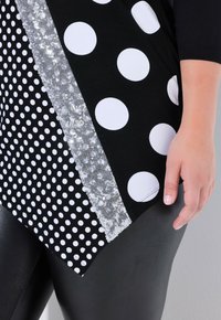 Černý top s různými vzory bílých puntíků, s postranním stříbrným flitrovým lemem. Materiál má jemnou strukturu.