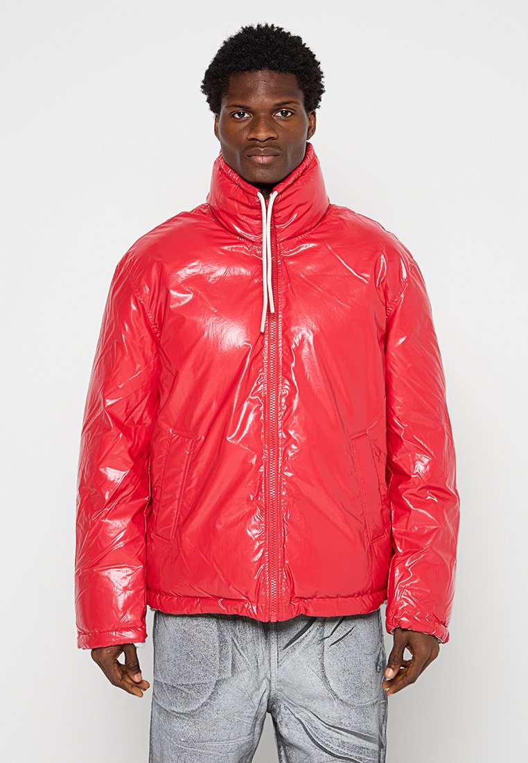 Chaqueta acolchada roja brillante con cuello alto, cierre frontal con cremallera y dos bolsillos laterales. Presenta puños elásticos y un ajuste holgado.