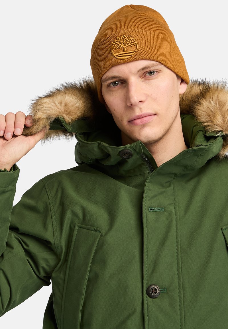 Parka verde con cappuccio foderato in pelliccia, chiusura a bottoni e tasche laterali. Accompagnata da un berretto lavorato a maglia marrone con logo in rilievo.