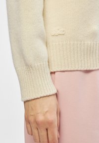 Κρέμα πουλόβερ πλεγμένο με ριμπ μανσέτες και στρίφωμα, με υφή και μια μικρή κεντητή λεπτομέρεια λογότυπου κοντά στο στρίφωμα.