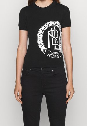 Kobieta ubrana w czarną bluzkę z krótkim rękawem z dużym białym, okrągłym logo „Lauren Ralph Lauren” oraz czarne spodnie.