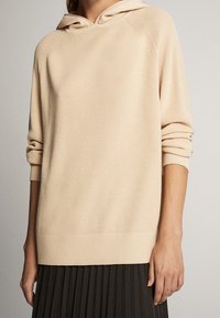 Pull à capuche beige clair avec un motif côtelé texturé. Comprend des manches larges et un ourlet arrondi, associé à une jupe plissée foncée.